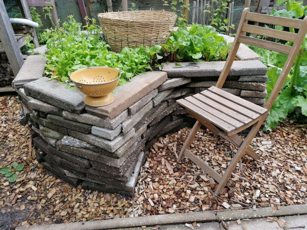 A round raised vegetable bed, with a height of 60 cm. Made of broken tiles. A chair is standing in front of the bed. Gardening can be done while sitting on the chair.